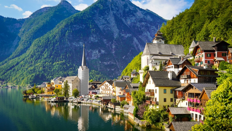 Avusturya Hallstatt'ta göl kıyısındaki ahşap evler, kilise kulesi ve arkadaki yeşil dağlar